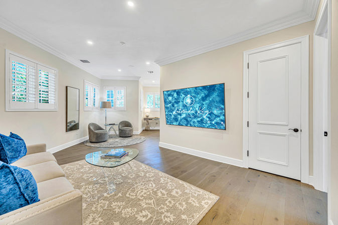 Bright modern living room/media space with wall-mounted TV showing blue ocean art, hardwood floors, glass coffee table, blue pillows and two grey accent chairs by shuttered windows.