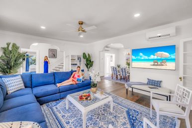 Sunny coastal-style living room with large blue sectional, patterned rug and white coffee table with fruit, wall TV showing a beach scene, and two women—one lounging with a tablet and one standing by the staircase; dining area visible in background.