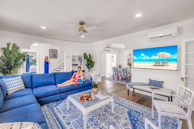 Sunny coastal-style living room with large blue sectional, patterned rug and white coffee table with fruit, wall TV showing a beach scene, and two women—one lounging with a tablet and one standing by the staircase; dining area visible in background.