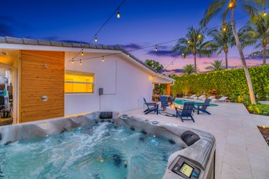 Evening tropical backyard oasis with a bubbling hot tub in the foreground, tiled poolside patio with lounge chairs around a fire pit, string lights overhead, palm trees and a purple sunset sky.