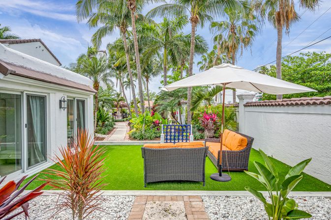 Sunny tropical backyard patio with tall palm trees, artificial turf, wicker seating with bright orange cushions under a white umbrella, sliding glass doors and a plant-lined pathway.