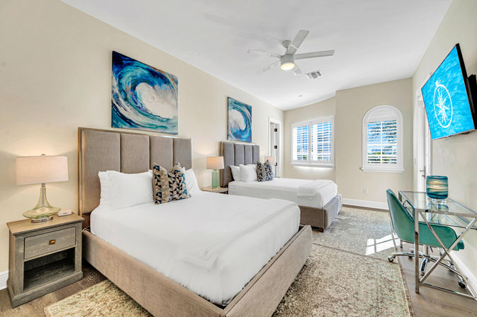 Sunlit coastal-style guest room with two queen beds, neutral upholstered headboards, ocean-wave artwork, bedside lamps, ceiling fan, wall-mounted TV, glass desk with teal chair, and plantation-shuttered windows.