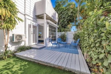 Sunny tropical backyard patio with white wooden deck, blue metal chairs and matching rug, sliding glass doors to a coastal-style home, surrounded by palm trees and green hedge