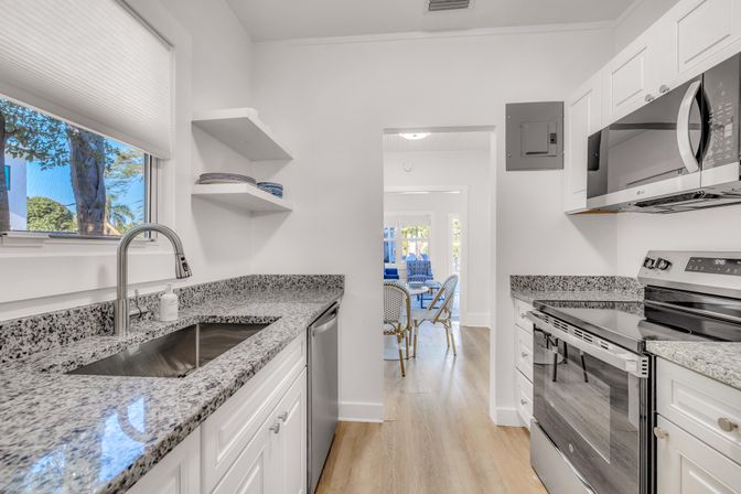 Sunlit modern white galley kitchen with speckled granite countertops, stainless steel sink and appliances, hardwood floors, and an open view to a blue-accented dining area.