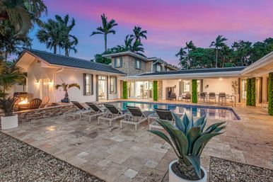 Luxury modern villa backyard with rectangular swimming pool, row of lounge chairs on stone tile patio, outdoor dining area and fire pit, agave planter and palm trees under a pink sunset sky