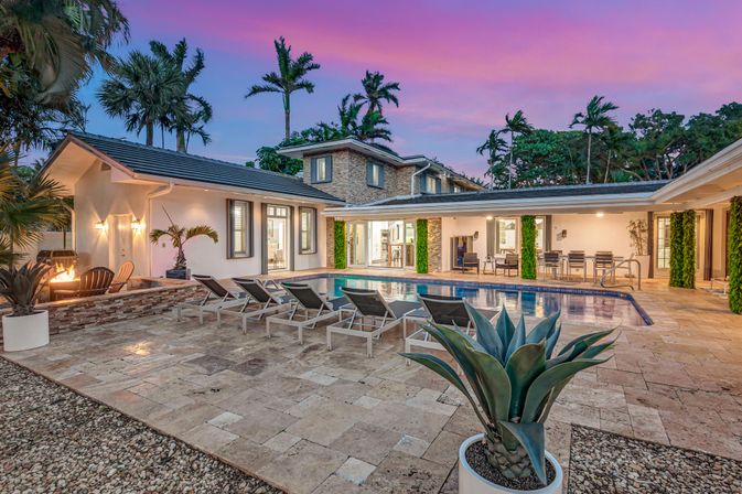 Luxury modern villa backyard with rectangular swimming pool, row of lounge chairs on stone tile patio, outdoor dining area and fire pit, agave planter and palm trees under a pink sunset sky