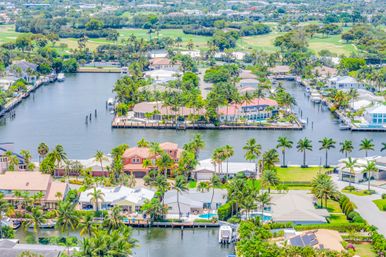 Aerial view of palm-lined waterfront homes and private docks along interconnecting canals with boats and a golf course in a sunny coastal Florida neighborhood.