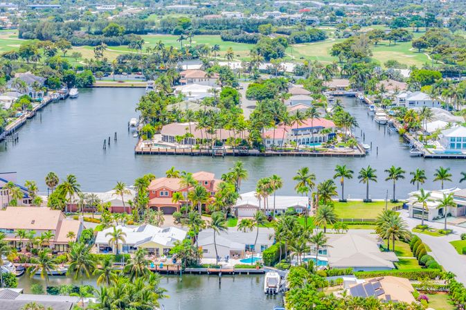 Aerial view of palm-lined waterfront homes and private docks along interconnecting canals with boats and a golf course in a sunny coastal Florida neighborhood.