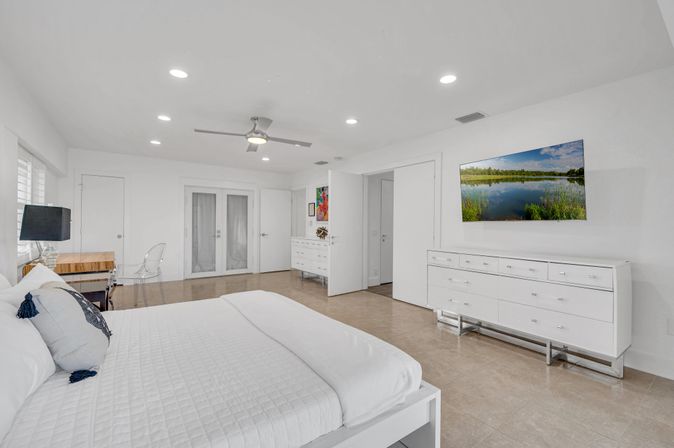 Bright modern master bedroom with white minimalist decor and airy coastal vibe, king bed with white quilt and navy tassel pillows, glass desk and acrylic chair, wall-mounted landscape TV above a white dresser, recessed lighting, ceiling fan, beige tile floors and double French doors