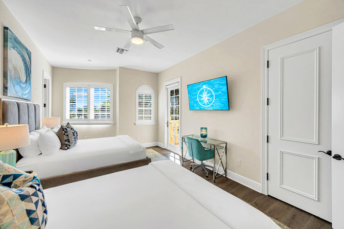 Bright coastal-style bedroom with two white beds and tufted headboards, wall-mounted TV, glass desk with teal chair, ceiling fan, plantation shutters and balcony door