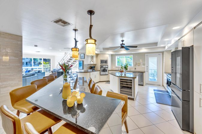 Bright modern open-concept kitchen — dark granite breakfast bar with tan leather stools and pendant lights, central island with built-in wine fridge, stainless appliances, white cabinets, tiled floor, ceiling fan, and open view to the living area and staircase