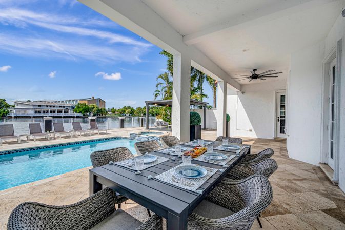 Waterfront poolside patio with covered dining table set for six, woven wicker chairs, loungers along a canal, palm trees and ceiling fan