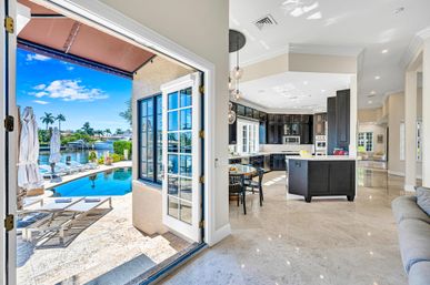 Sun-drenched luxury waterfront home seen through open French doors to a poolside patio and canal marina; open-plan kitchen with dark cabinets, island, dining area and marble floors, palm trees and boats in the background.