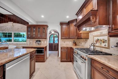 Spacious luxury kitchen with dark cherry wood cabinets, granite island and countertops, stainless steel gas range, decorative tile backsplash, arched doorway and window with pool view.