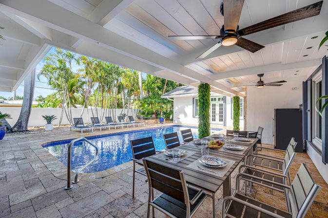 Covered poolside patio with large outdoor dining table set for a meal, ceiling fans, blue-tiled lap pool, sun loungers and palm trees in a tropical backyard.