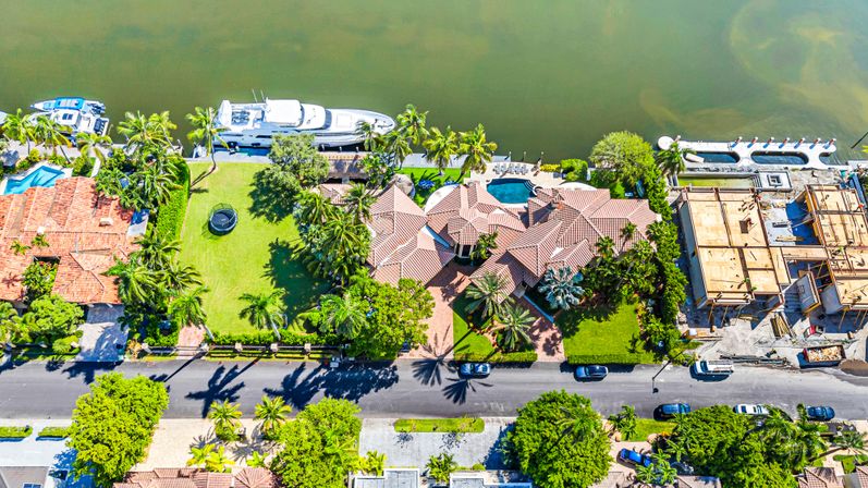 Aerial drone view of a sunny tropical waterfront estate with red-tile roof, palm-lined lawn, backyard pool and a luxury yacht docked along a calm canal.