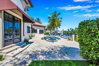 Sunny waterfront Mediterranean-style villa patio with circular spa and pool, palm trees, lounge chairs and private dock with a boat under a bright blue sky