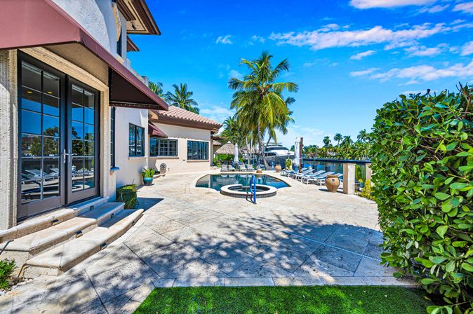 Sunny waterfront Mediterranean-style villa patio with circular spa and pool, palm trees, lounge chairs and private dock with a boat under a bright blue sky