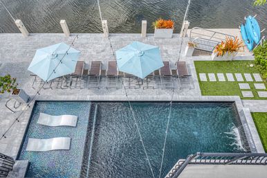 Aerial view of a modern waterfront pool deck with two light-blue umbrellas over lounge chairs, mosaic sun shelves with in-pool loungers, string lights, stepping stones and stacked paddleboards along a canal.