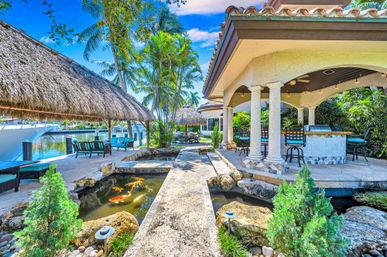 Tropical waterfront patio with stone walkway, thatched tiki huts, koi pond with colorful fish, outdoor tiled bar and stools, palm trees and docked yacht.