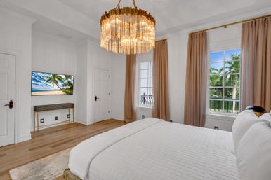 Bright luxury master bedroom with white quilted bed, gold crystal chandelier, hardwood floors, wall-mounted TV showing a beach scene, and tall beige curtains framing windows with a tropical palm-tree view.