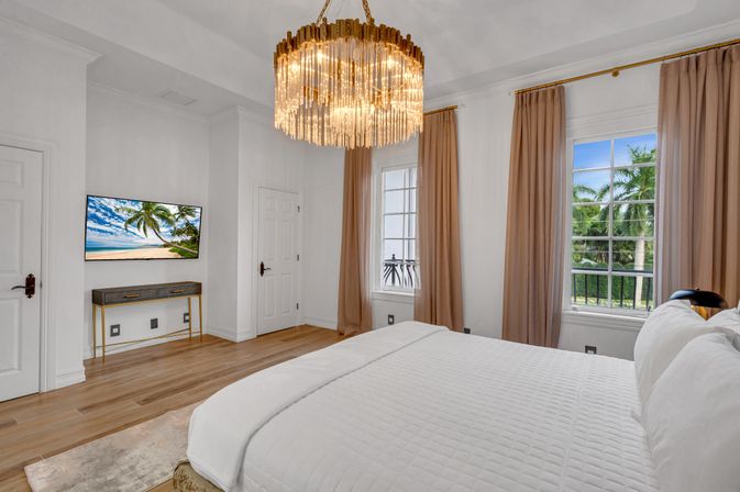 Bright luxury master bedroom with white quilted bed, gold crystal chandelier, hardwood floors, wall-mounted TV showing a beach scene, and tall beige curtains framing windows with a tropical palm-tree view.