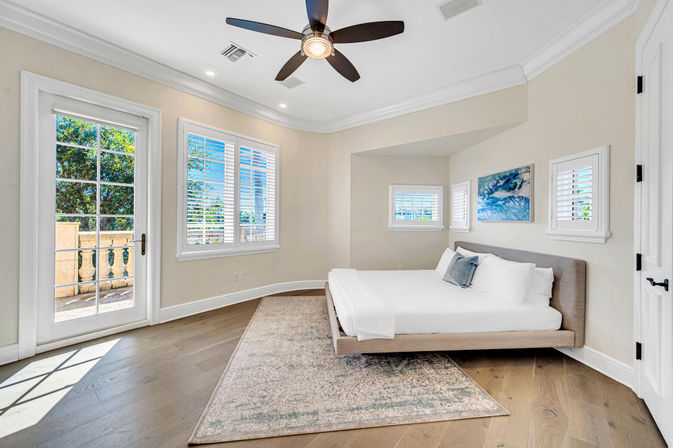 Airy sunlit master bedroom with low platform bed, hardwood floors, ceiling fan, plantation shutters, abstract blue wall art and French door opening to a balcony with greenery.