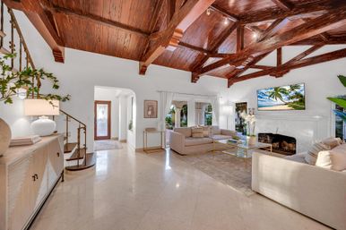 Sunlit Mediterranean-style living room with exposed dark wood beam ceiling, polished marble floors, neutral sofas around a glass coffee table, white fireplace with wall-mounted TV, spiral iron staircase and arched doorway opening to a garden.