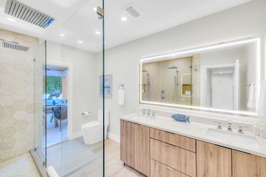 Bright modern coastal bathroom with frameless glass rain shower and hex tile, dual-sink wood vanity with backlit mirror and white countertops, toilet and doorway opening to a sunlit patio