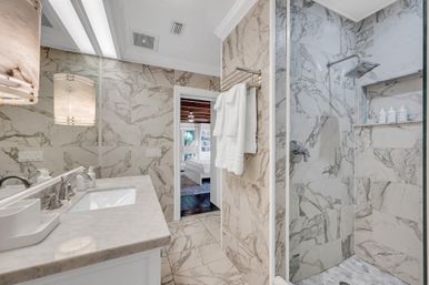 Spa-like luxury marble bathroom in a modern home featuring veined marble walls and floor, marble vanity with sink and mirror, glass walk-in shower with rainfall head and built-in shelf, chrome fixtures and towel rack, doorway opening to bedroom.