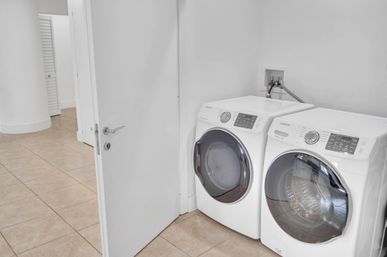 Bright modern laundry nook with matching white front-loading washer and dryer on beige tile, open white door and visible water hookups.