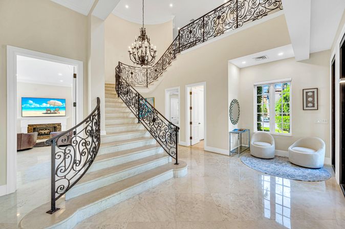 Grand sunlit foyer in a luxury home with a sweeping wrought-iron staircase, polished marble floors, crystal chandelier and cozy window seating with two rounded chairs.