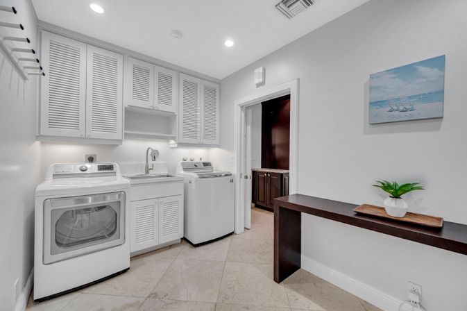 Bright modern laundry room with white washer and dryer, utility sink, louvered upper cabinets, tile floor, dark folding table with potted plant and beach wall art.