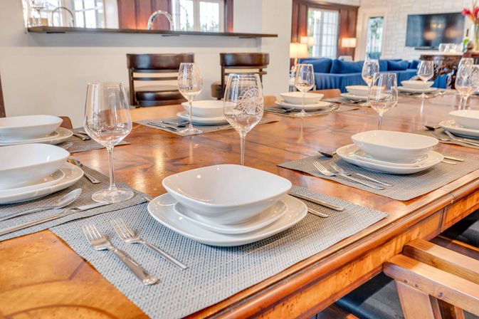 Long wooden dining table in an open-concept home set for a meal with white modern dinnerware, wine glasses, gray woven placemats and stainless flatware, kitchen pass-through and blue sofa in the background.