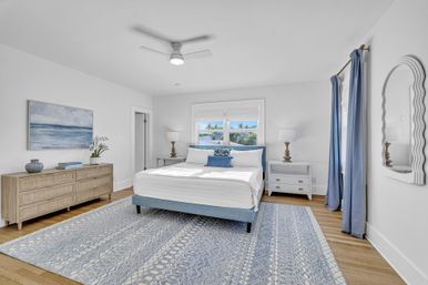 Bright coastal-style master bedroom with a blue upholstered bed and white bedding, patterned blue area rug on light wood floors, matching nightstands and lamps, dresser with ocean artwork, large window with blue curtains and ceiling fan.