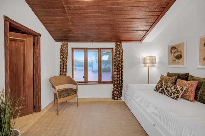 Cozy sunlit bedroom with a wood-paneled ceiling, wicker chair by a triple window with patterned curtains, white daybed piled with decorative pillows, floor lamp, and a patio window revealing a water view.