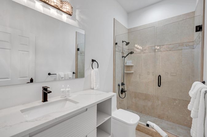 Modern master bathroom with white marble vanity, black faucet, large mirror, and glass-enclosed beige-tile shower with dual showerheads and plush towels.