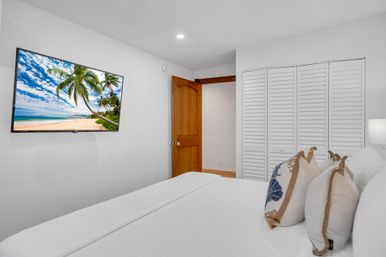 Cozy coastal bedroom with white linens and decorative pillows, wall-mounted TV showing a palm-tree beach, wooden door and white louvered closet.
