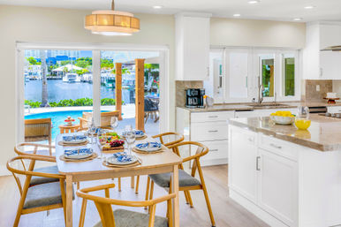 Bright modern coastal waterfront kitchen and dining area — wooden table set for six, white marble island, sliding doors opening to a pool and marina with boats.