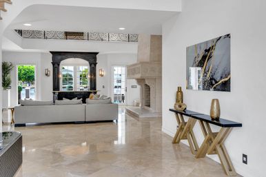 Sunlit luxury waterfront living room with glossy marble floors, pale gray sectional sofa facing a dark wooden bar and carved stone fireplace; abstract gold-and-gray wall art and black console table with gold vases.