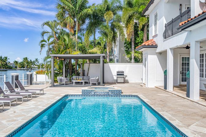 Sunny waterfront backyard with turquoise pool, mosaic-tiled spa, shaded cabana lounge, chaise lounges, barbecue grill and palm trees beside a white coastal home.