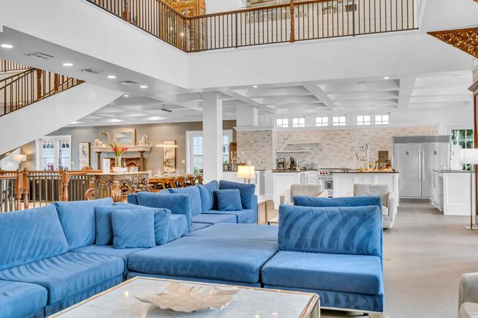 Airy two-story open-concept living room and kitchen with a plush blue sectional, marble coffee table, white island kitchen with stainless steel refrigerator, dining table, staircase and wooden balcony railing.