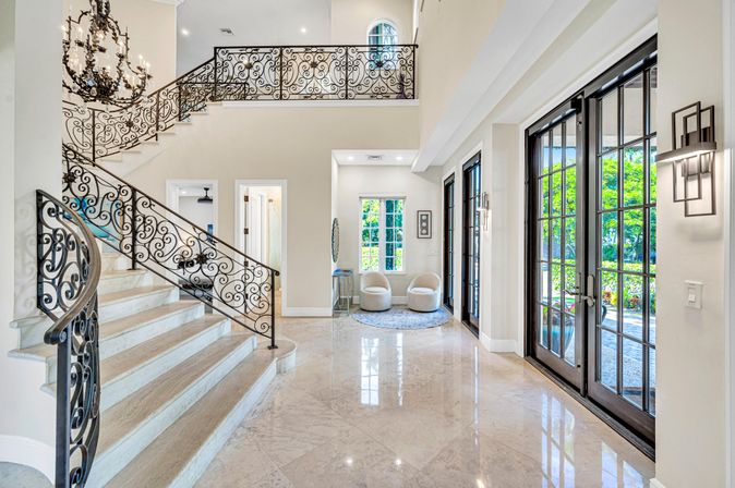 Sunlit luxury foyer with polished marble floors, curved grand staircase and ornate wrought-iron railing, chandelier, cozy seating nook and tall French doors opening to a garden