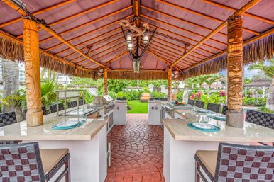 Tropical tiki-style outdoor kitchen and bar under a thatched palapa with carved wooden pillars, marble countertops, built-in stainless steel grill, bar seating, and lush backyard garden.