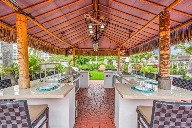 Tropical tiki-style outdoor kitchen and bar under a thatched palapa with carved wooden pillars, marble countertops, built-in stainless steel grill, bar seating, and lush backyard garden.