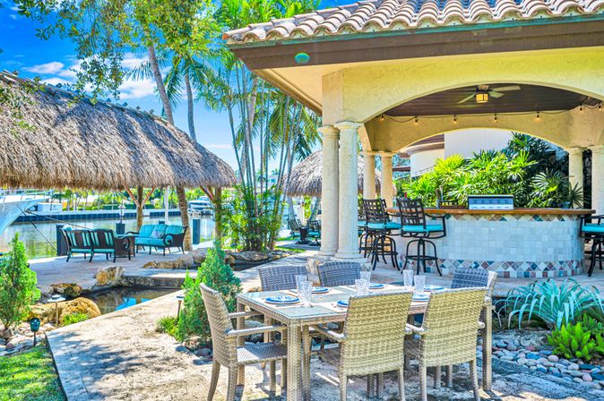 Tropical waterfront patio: wicker dining table for six, tiled outdoor bar with stools under a covered terrace, thatched tiki lounges by the marina and palm trees.