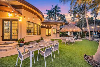 Tropical sunset backyard patio with modern outdoor dining tables, hanging string lights, thatched tiki huts and palm trees on a manicured lawn
