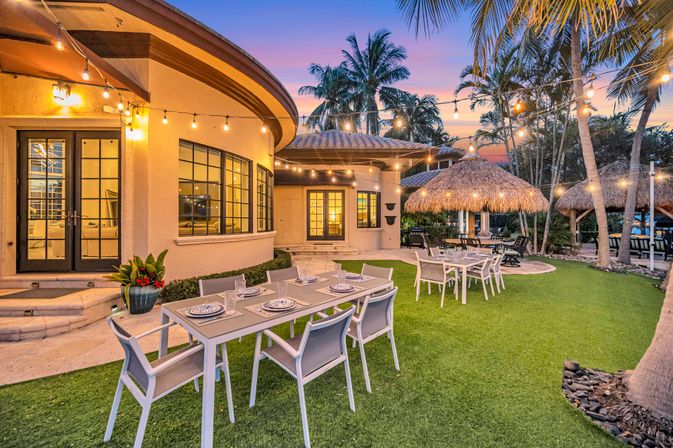 Tropical sunset backyard patio with modern outdoor dining tables, hanging string lights, thatched tiki huts and palm trees on a manicured lawn
