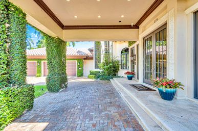 Sunny Mediterranean-style home entrance with ivy-covered porte-cochere, brick paver driveway, tiled roof, double glass French doors, garage doors, and blue planters with tropical foliage.