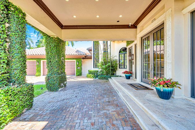 Sunny Mediterranean-style home entrance with ivy-covered porte-cochere, brick paver driveway, tiled roof, double glass French doors, garage doors, and blue planters with tropical foliage.